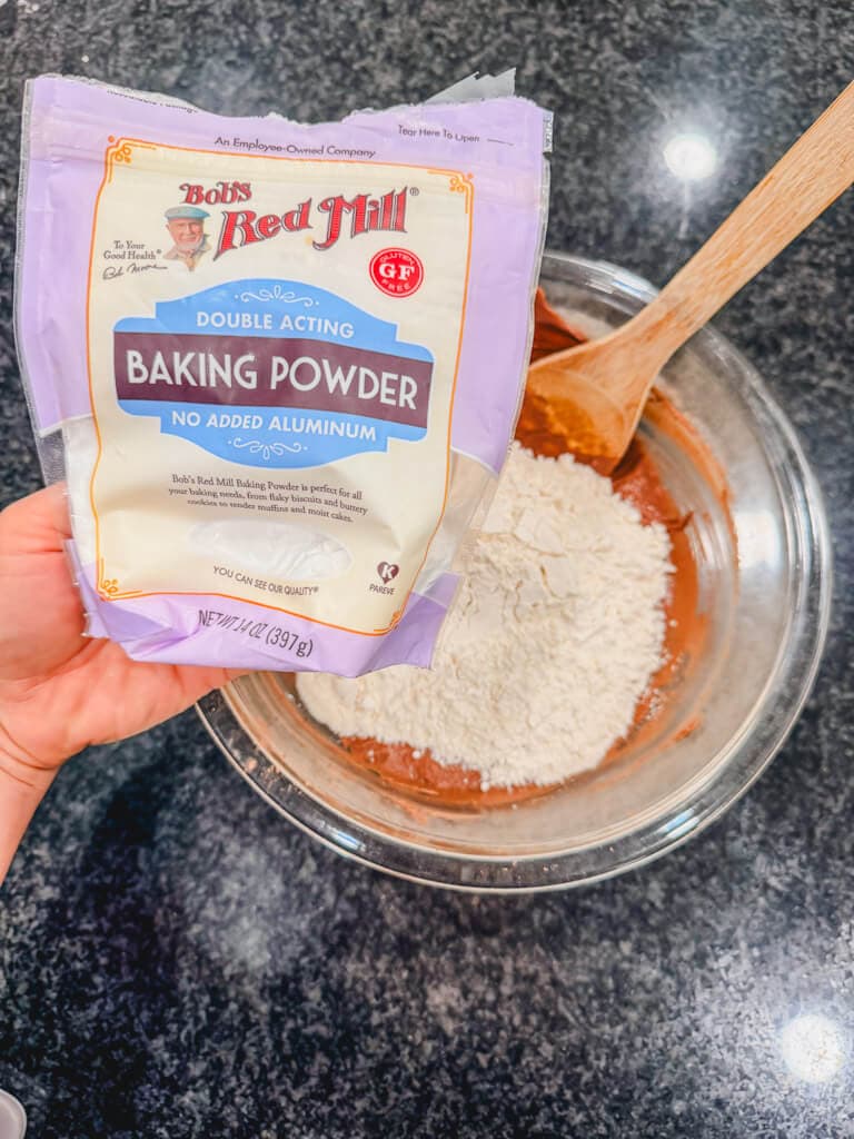 A bag of baking powder being held over the mixing bowl for chocolate crinkle cookies.