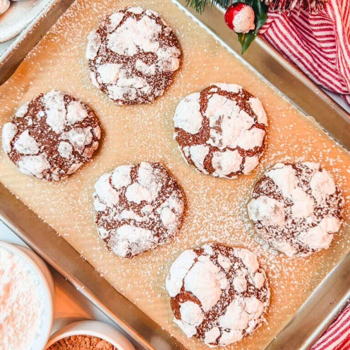 close up of chocolate crinkle cookies on a baking sheet next to a bowl of powdered sugar and a bolw of cocoa powder.