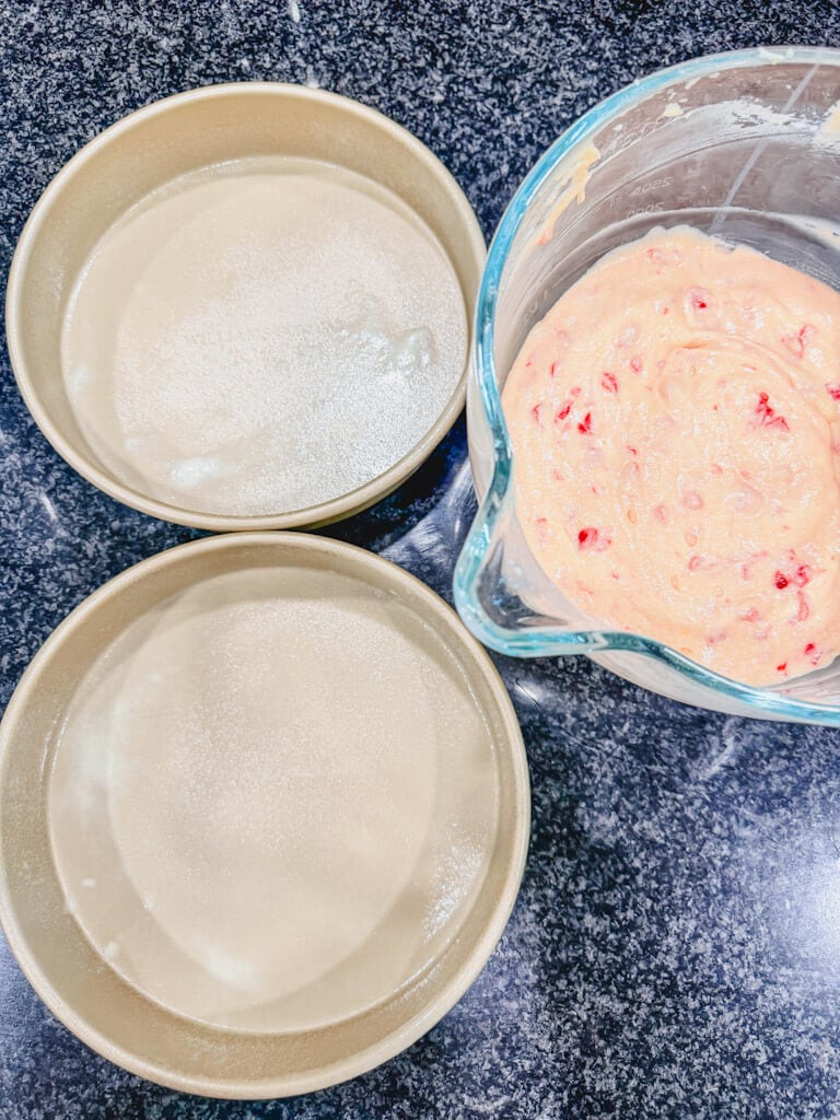 the cake batter sitting next to two cake pans that have been sprayed with nonstick spray.