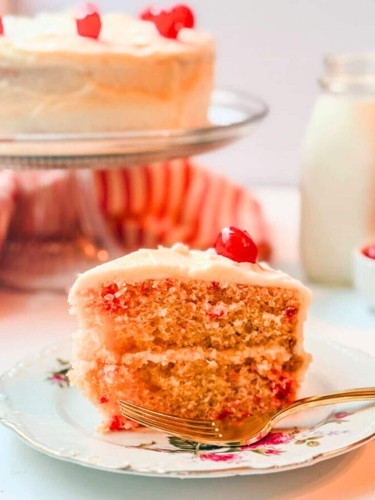 a slice of cherry chip cake on a plate with the cake and a bottle of milk in the background.