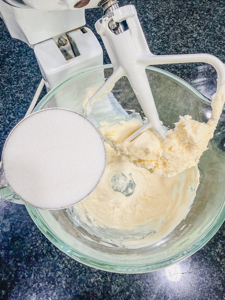 a cup of sugar being held over the creamed butter in the mixing bowl.