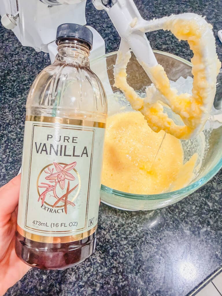 A bottle of vanilla extract being held over the bowl of butter, sugar, and eggs.