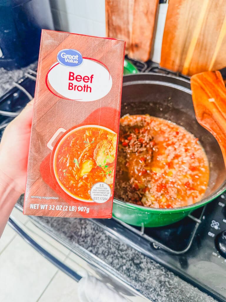 A carton of beef broth being held over the large pot.