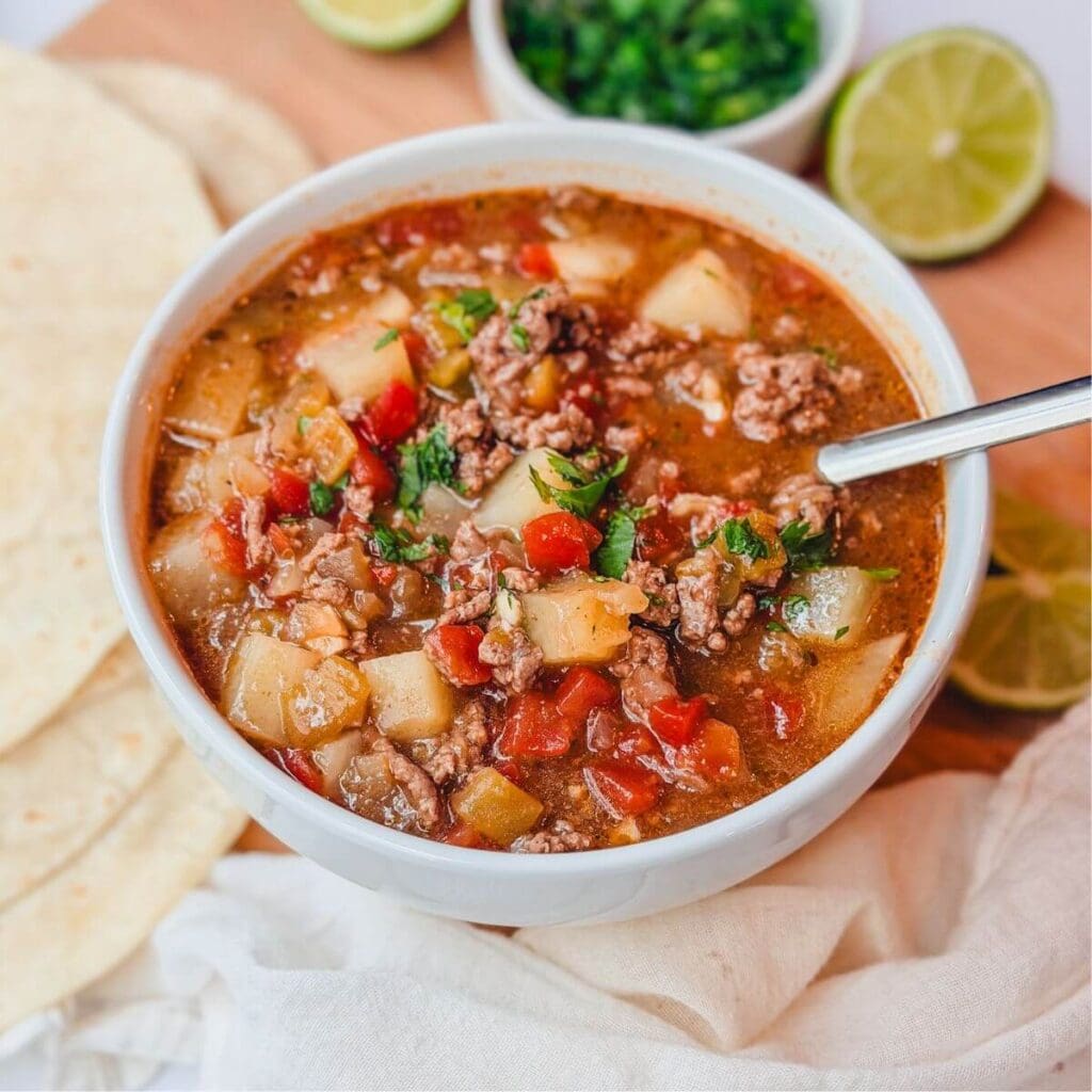 Ground beef chile stew in a white bowl next to some limes, flour tortillas, and chopped cilantro,