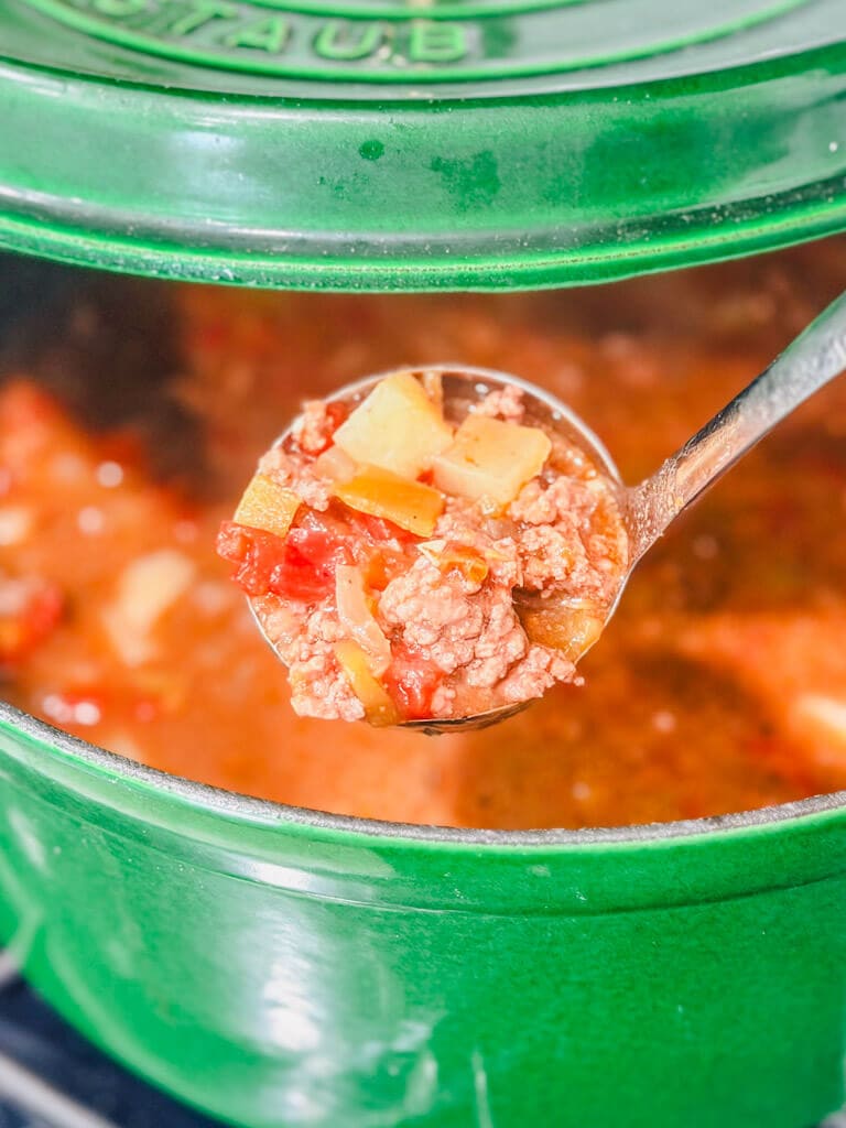 close up of a ladle with a serving of ground beef chile stew in a green Dutch oven.