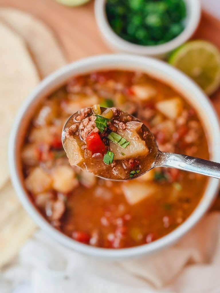 a spoonful of ground beef chile stew being held over a soup bowl.