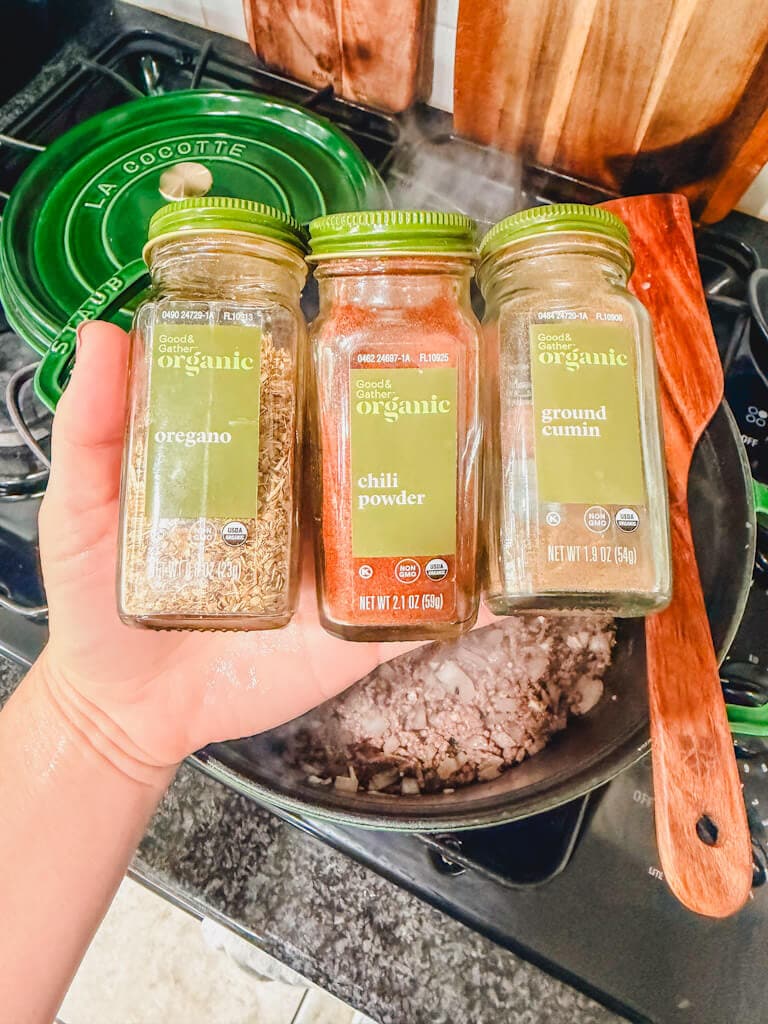 Three seasoning jars being held over the pot of ground beef.
