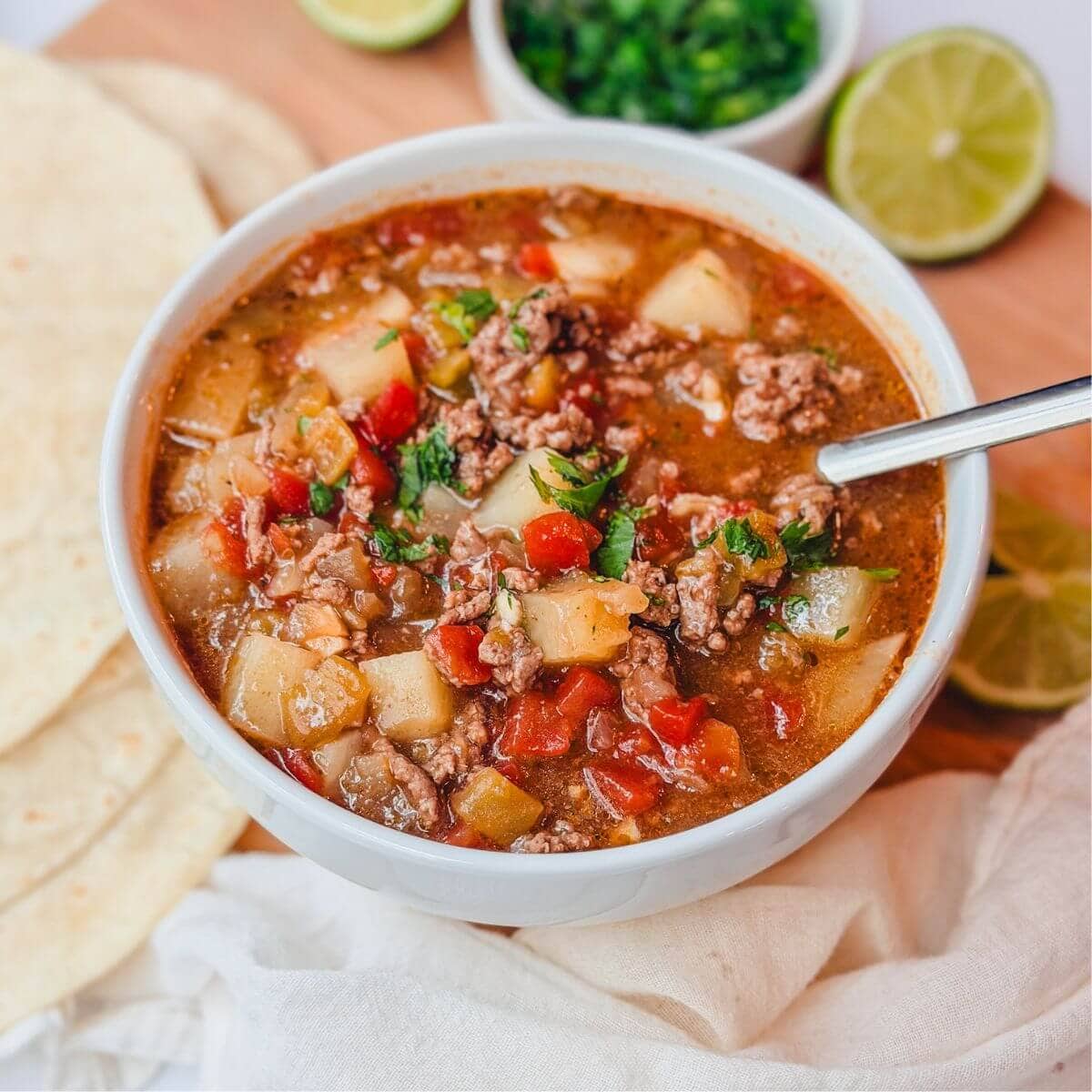 Ground beef chile stew in a white bowl next to some limes, flour tortillas, and chopped cilantro,