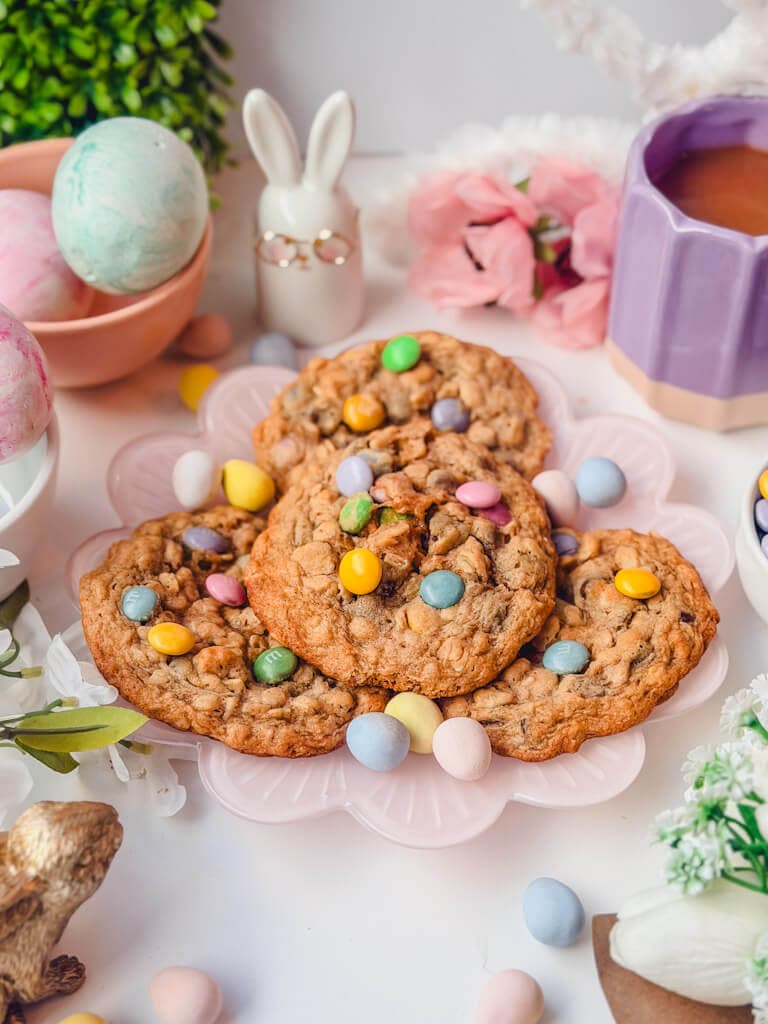 Four monster cookies with M&M's on a pink plate next to Easter decor props.
