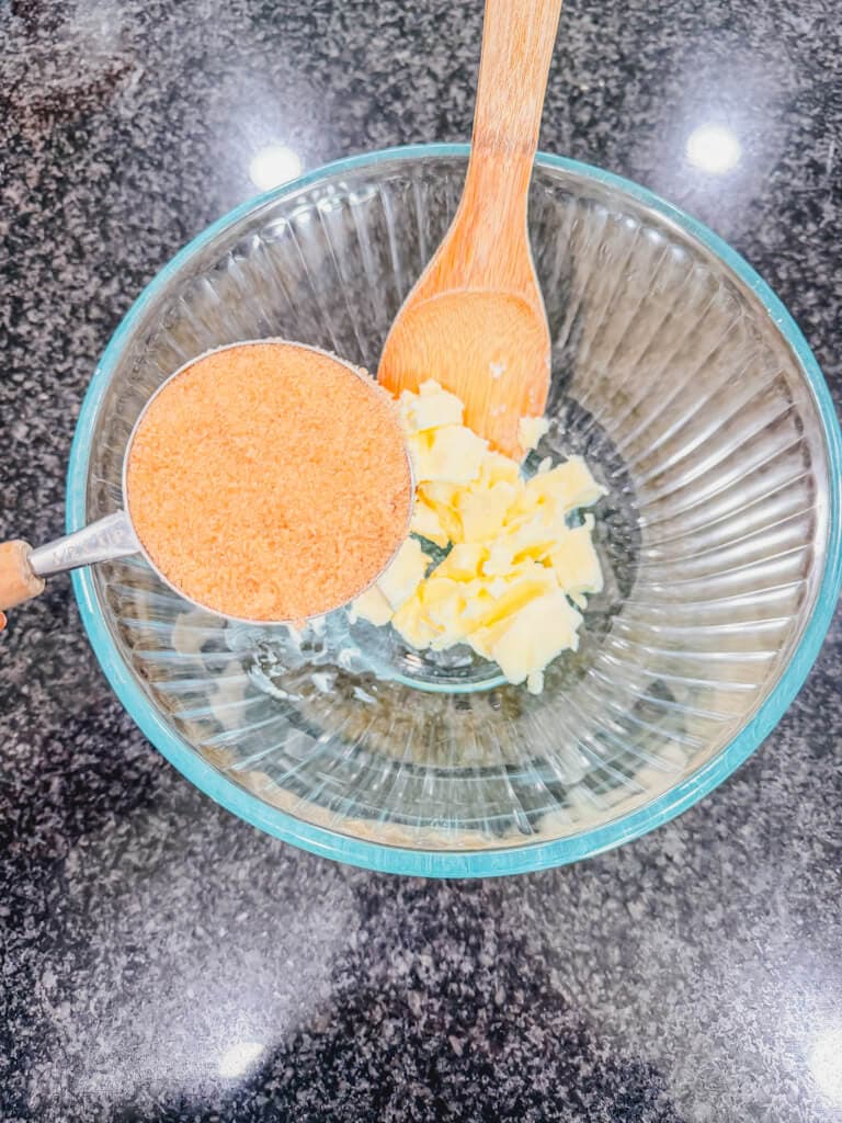 A glass mixing bowl with softened butter and a measuring cup filled with brown sugar bean held over the bowl.