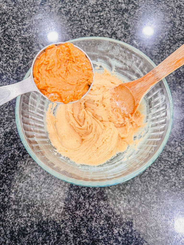 A measuring cup filled with peanut butter being held over the bowl of cookie dough.