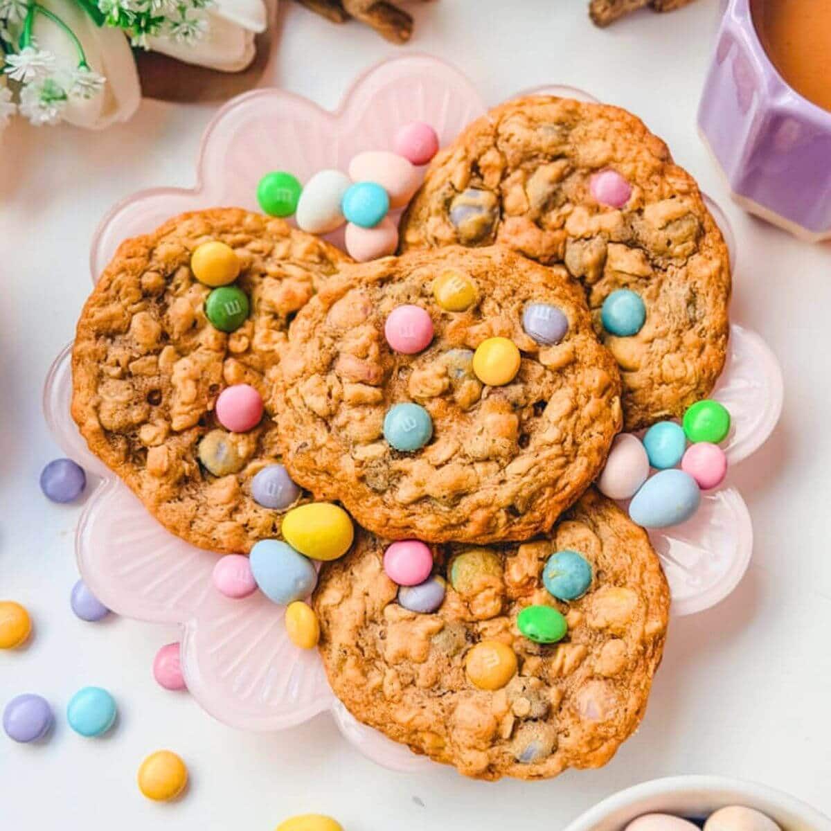 Four Monster Cookies with M&M's on a pastel pink plate next to a mug of coffee and white tulips.
