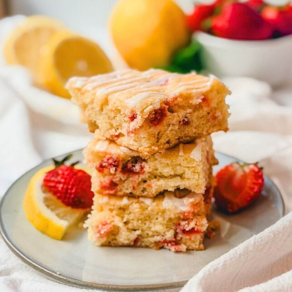 Close up of 3 strawberry lemon bars stacked on a plate with lemons and strawberries blurred out in the background.