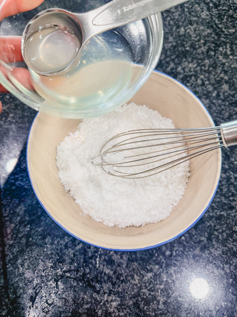 A small bowl filled with powdered sugar with a small bowl of lemon juice being held over the bowl.