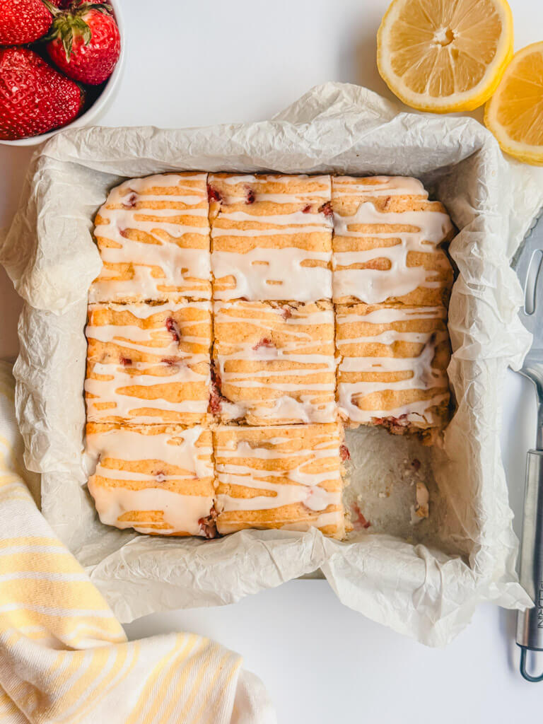 overhead view of strawberry lemon bars sliced into 9 bars in a baking dish with strawberries and lemons next to the dish. 