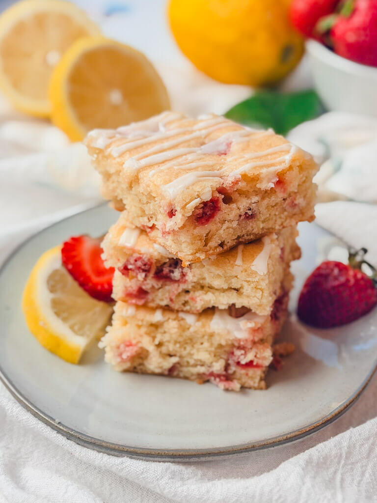 Three strawberry lemon bars stacked on top of each other on a plate with lemons and strawberries in the background.