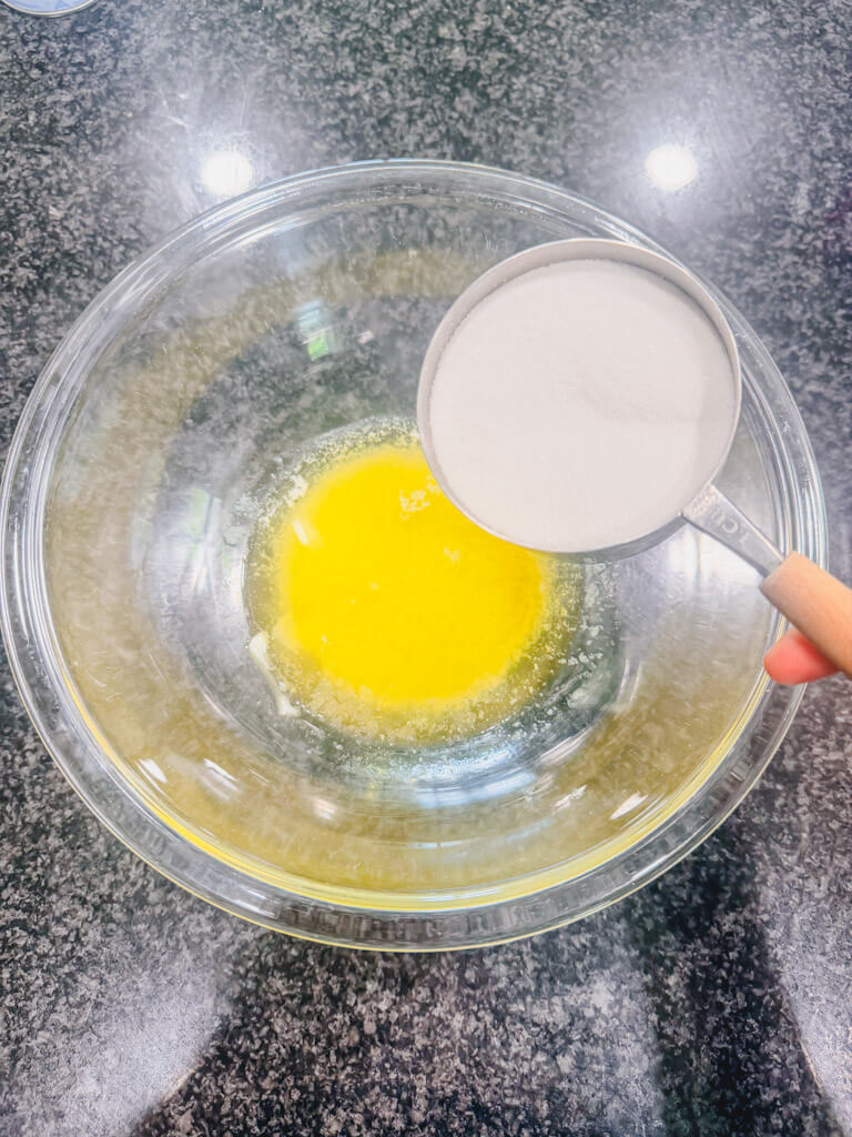 Measuring cup with sugar being held over the bowl of melted butter.