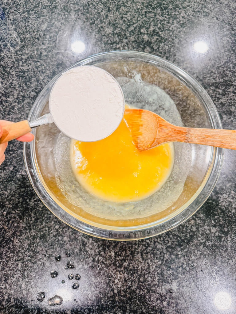 A measuring cup, holding flower being held over the mixing bowl of wood ingredients for strawberry lemon bars.