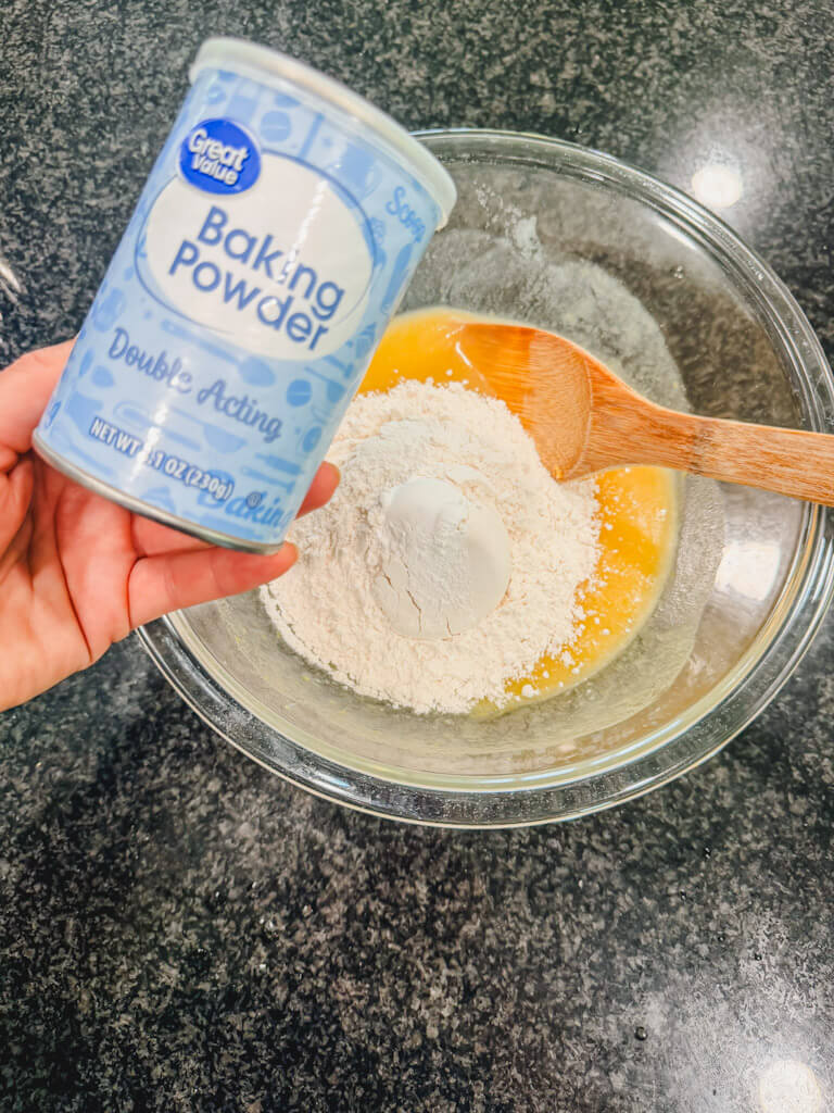 A hand holding a jar of baking powder over the mixing bowl.