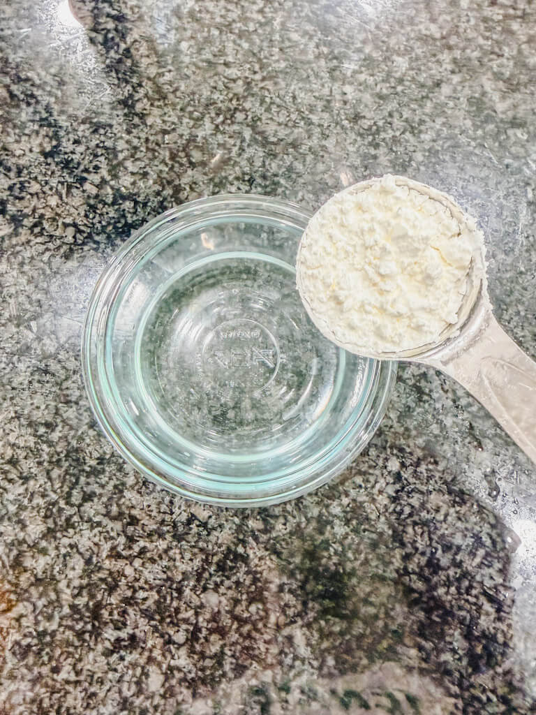 a tablespoon of cornstarch being held over a small bowl of water to make a cornstarch slurry.