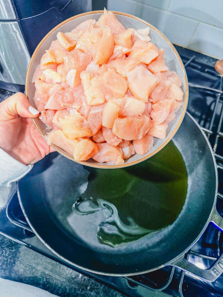 a bowl of cubed chicken breasts being held over a skillet that has warm olive oil.