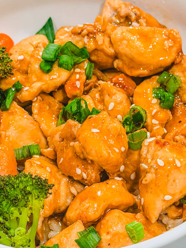 close up of teriyaki chicken in a bowl that is topped with sesame seeds and green onions.