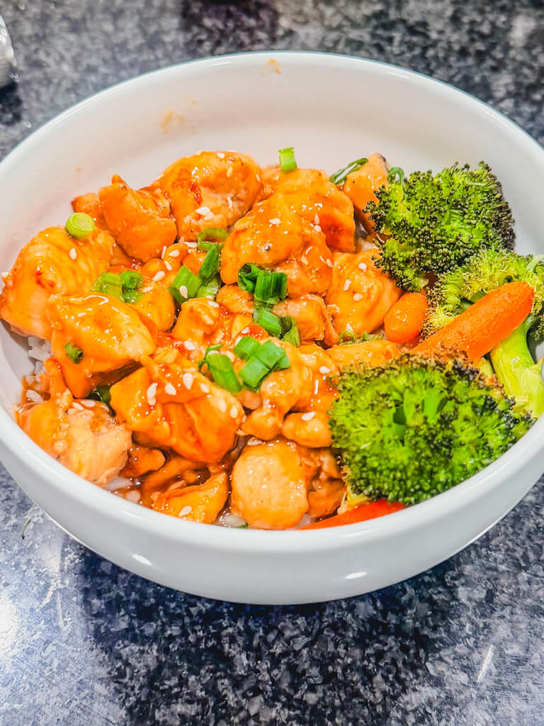 Teriyaki chicken in a bowl with roasted veggies and topped with sesame seeds and green onions.
