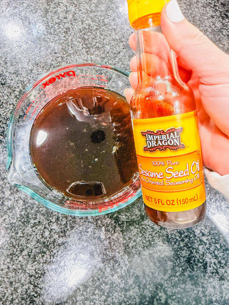 A hand holding a bottle of Sesame seed oil about to be added to the measuring cup.