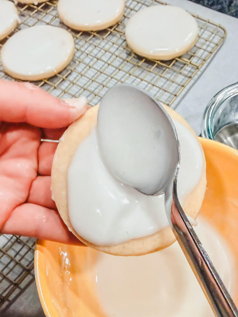 A hand holding a cookie and a spoon is spooning the glaze over the top of the cookie.