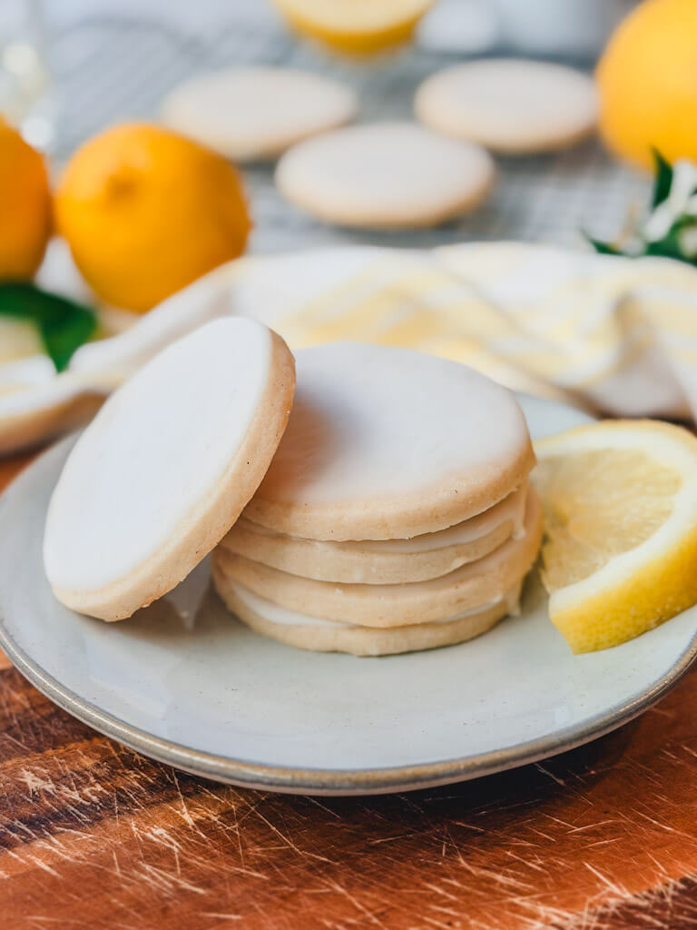Stack of lemon vanilla bean shortbread cookies on a ceramic plate with lemon slices and glazed cookies in the background.