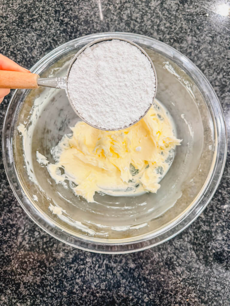 A measuring cup, holding powdered sugar over the softened butter in the mixing bowl.
