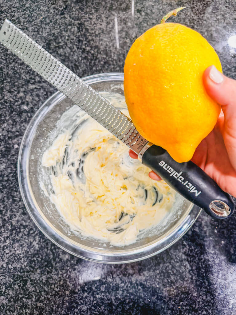 A lemon and a zester being held over the bowl of softened butter and powdered sugar.