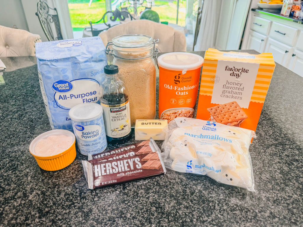 Ingredients for Cast Iron Skillet S'mores Pie including flour, oats, graham crackers, marshmallows, chocolate, butter, brown sugar, and vanilla extract displayed on a kitchen counter.