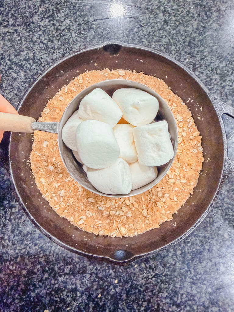 Large marshmallows being measured over the graham cracker crust in a cast iron skillet.