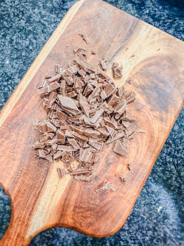 Milk chocolate bars coarsely chopped on a wooden cutting board.