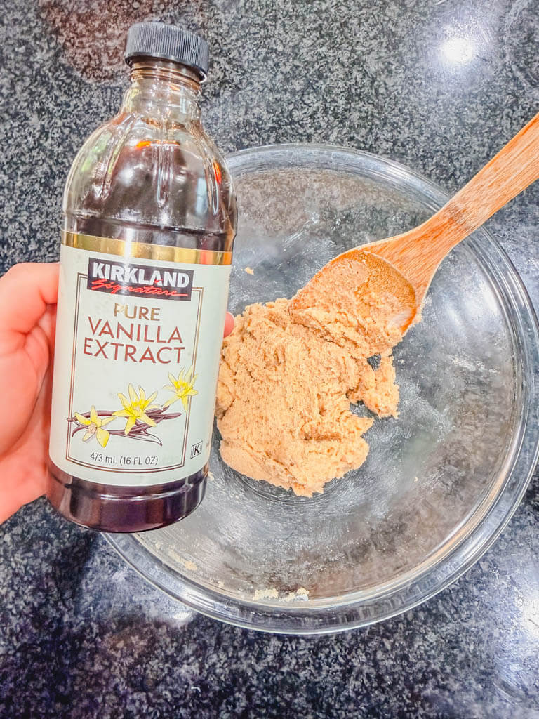 Vanilla extract bottle being held over the creamed butter and brown sugar mixture in a glass mixing bowl.