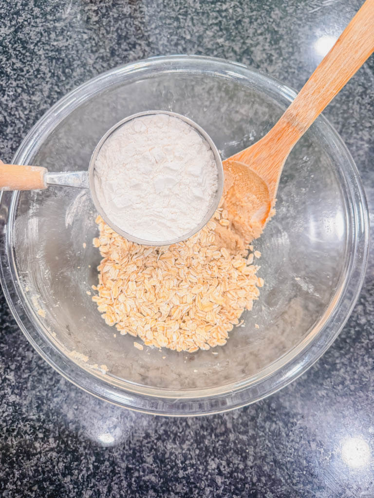 All-purpose flour being added to the butter, brown sugar, and oats mixture in a glass mixing bowl.