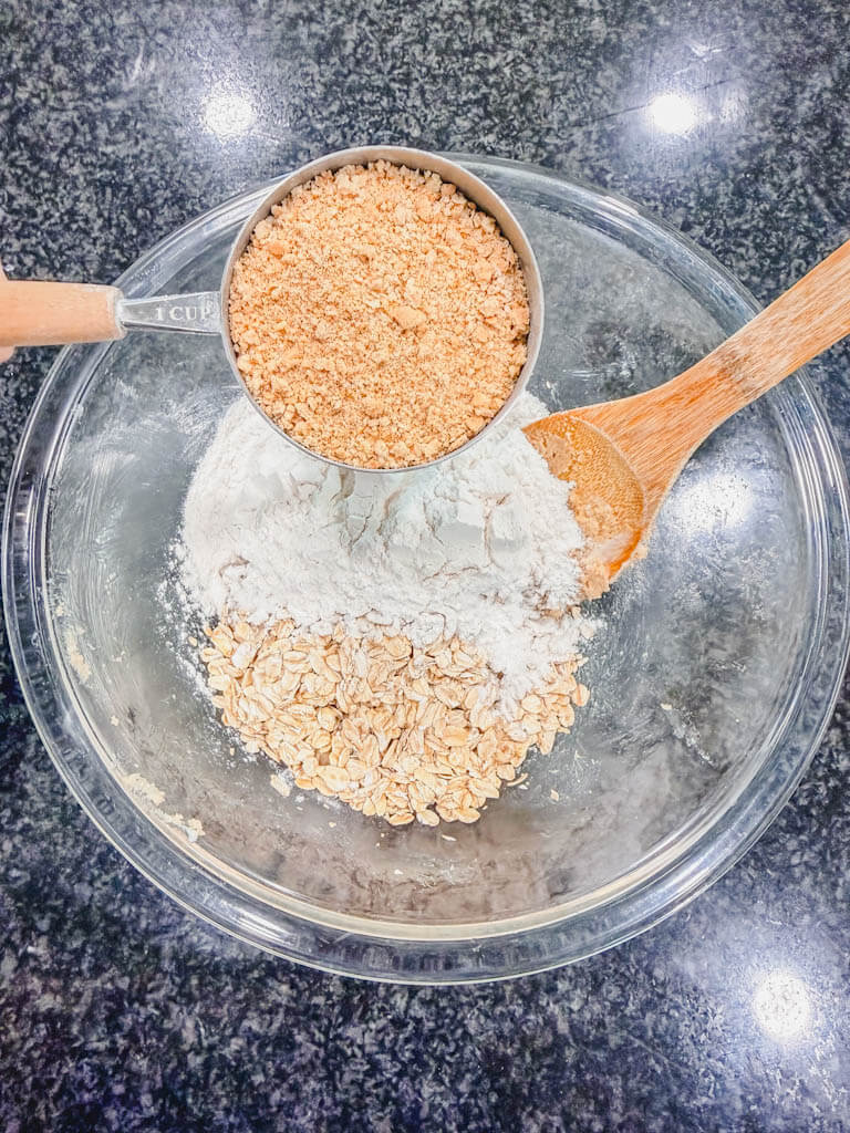 Graham cracker crumbs being added to oats and flour in a glass mixing bowl.