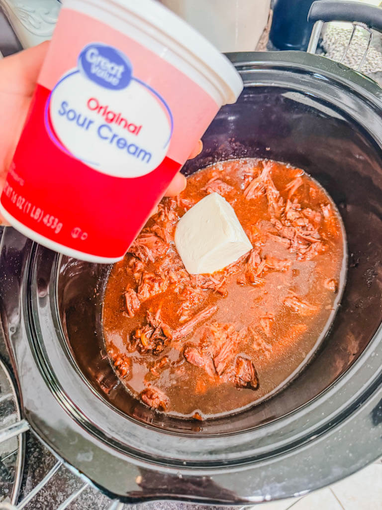 a small carton of sour cream being held over the slow cooker.
