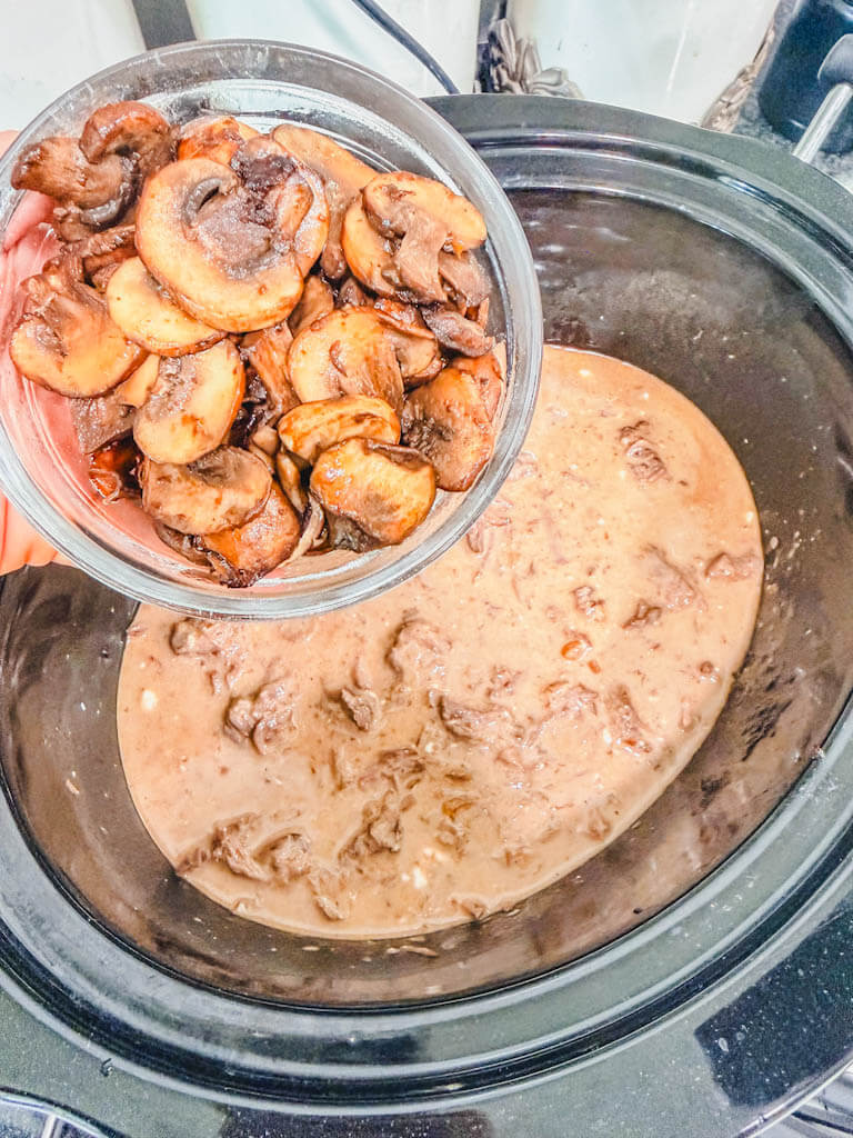 a small bowl of sauteed mushrooms being held over the slow cooker about to be added to the beef.