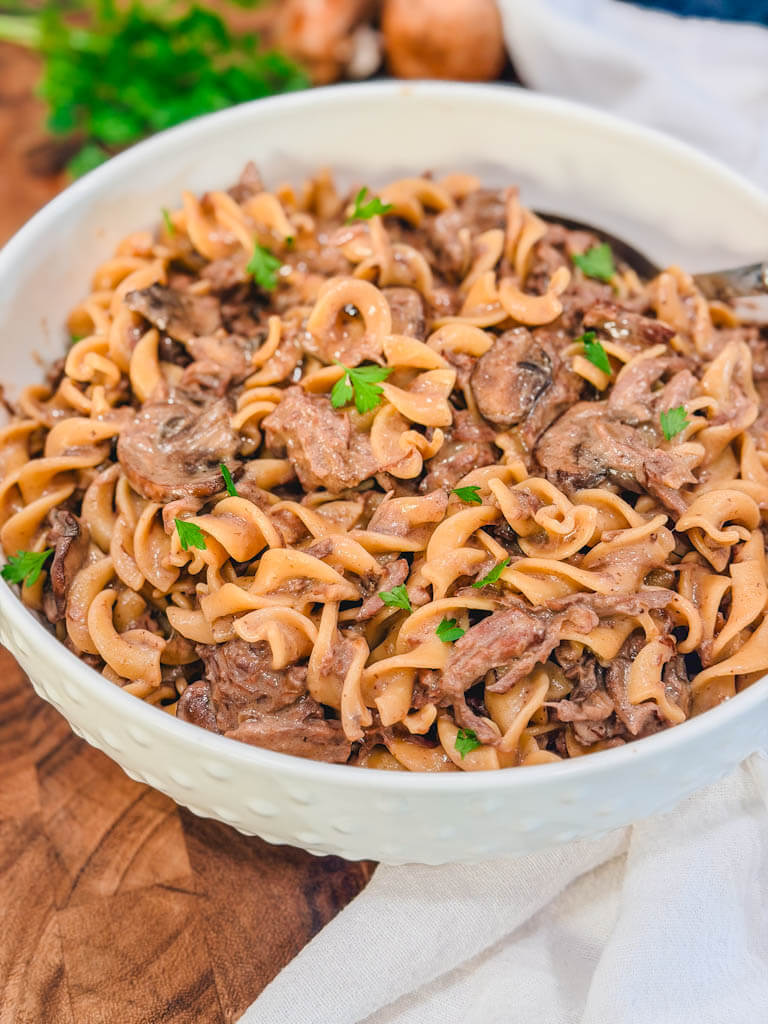 slow cooker beef stroganoff in a white bowl with parsley and mushrooms in the background.