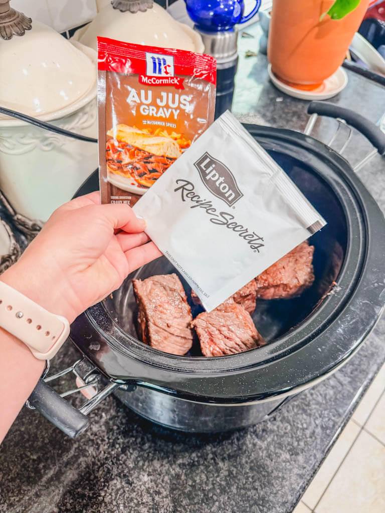 a packet of au jus and onion soup mix being held over the slow cooker.
