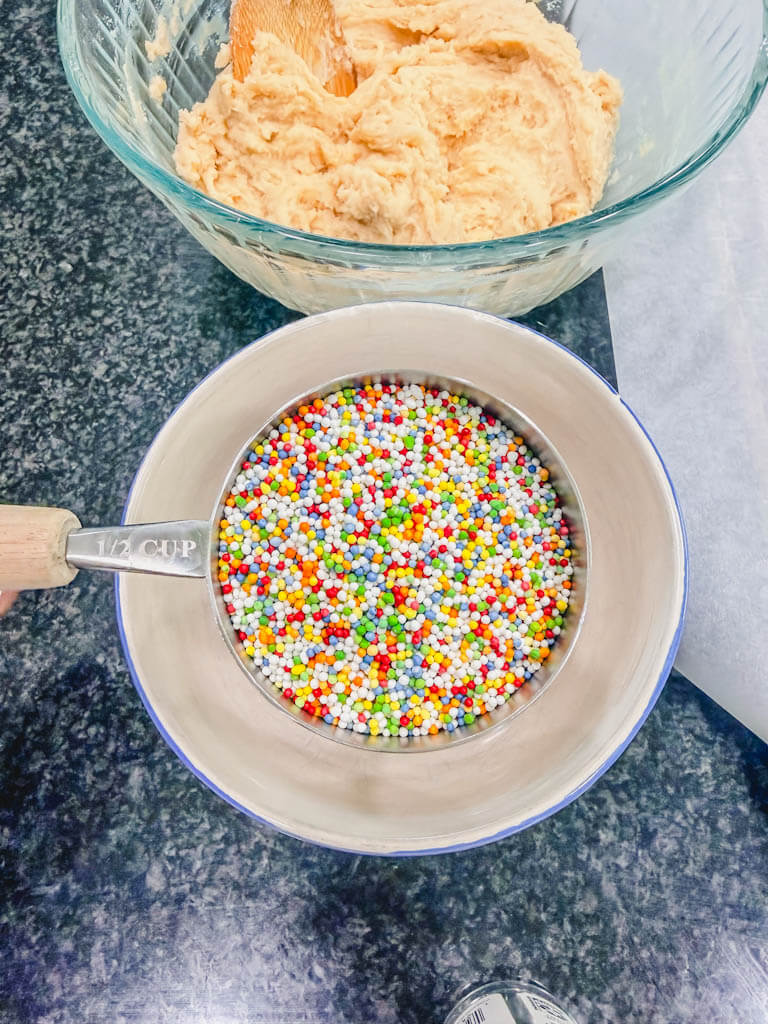 a measuring cup holding nonpareil rainbow sprinkles over an empty bowl, with the cookie dough in the background.
