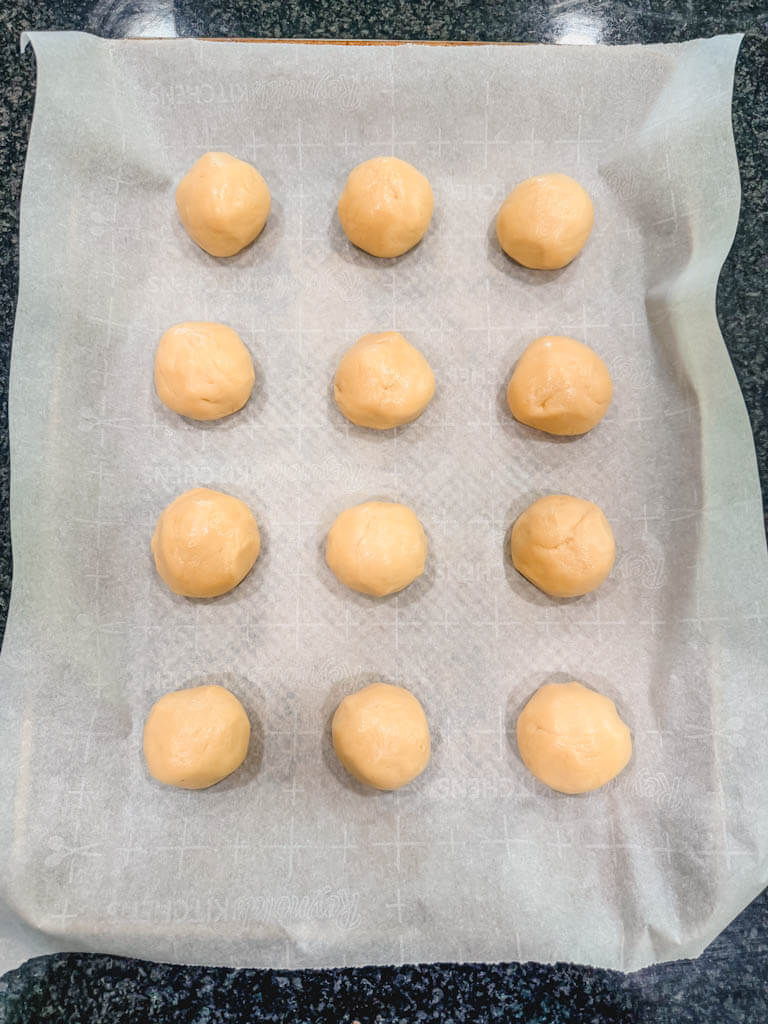 Twelve cookie dough balls resting on parchment paper on a cookie sheet.