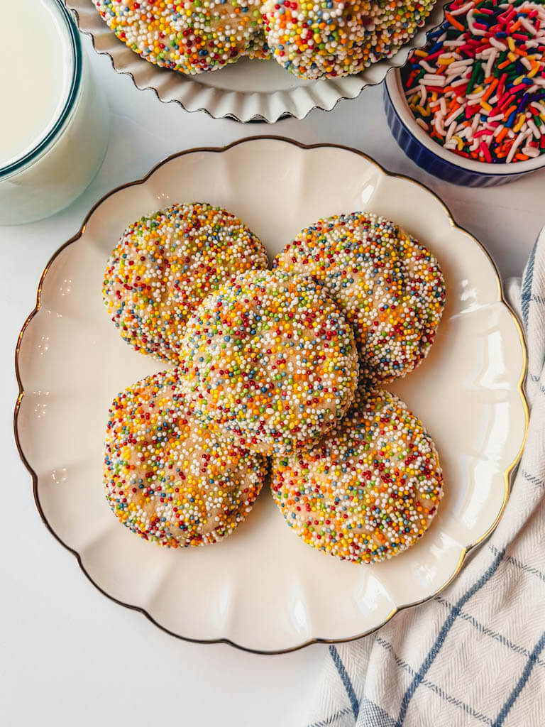 overhead view of sprinkle sugar cookies on a plate with a glass of milk and a small bowl of sprinkles next to the plate.