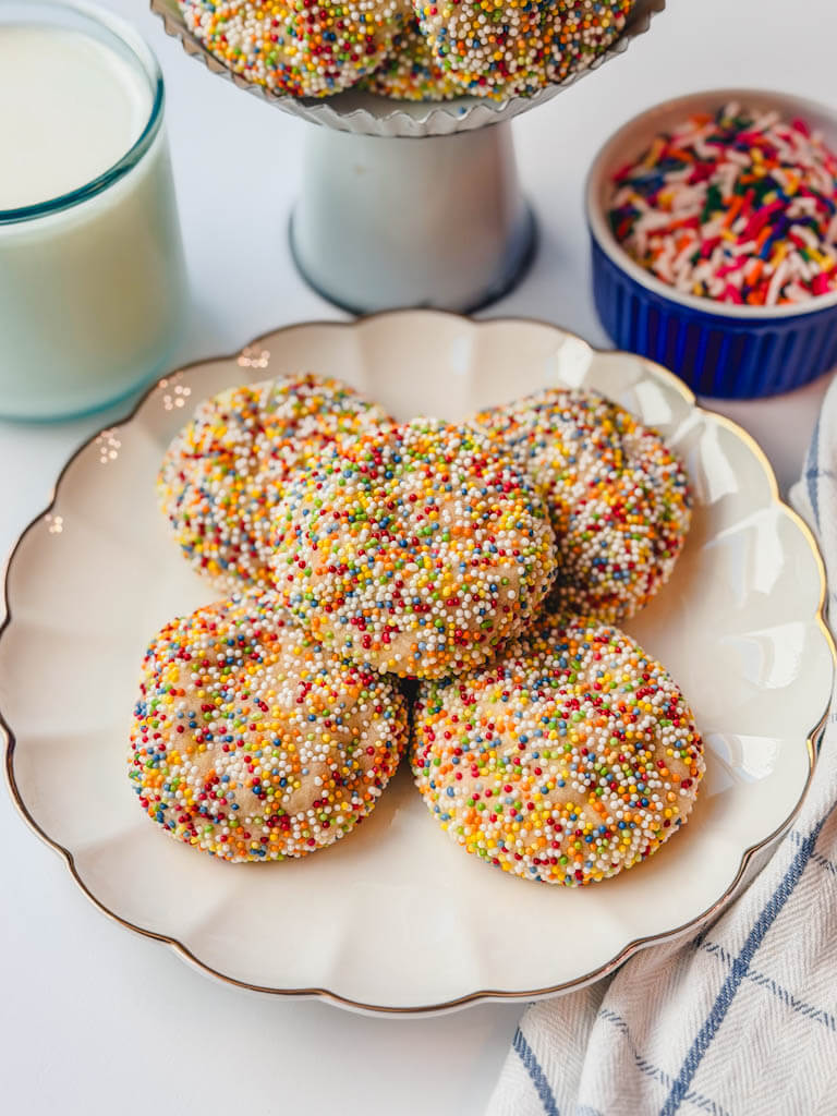 side angle view of sprinkle sugar cookies on a white scalloped plate with more cookies on a white stand behind the plate.