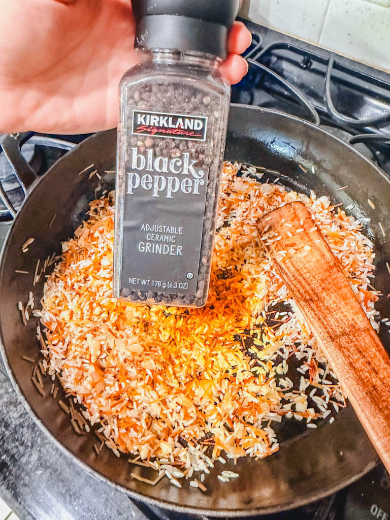 A bottle of black pepper being held over the skillet.