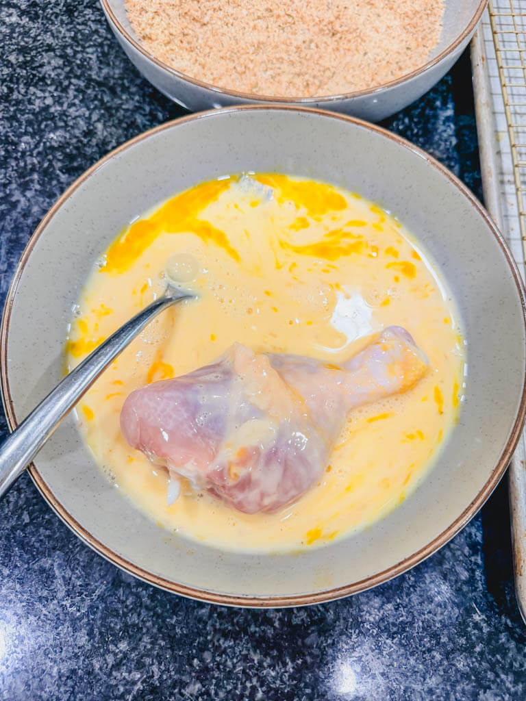 Chicken drumstick dipped into the egg wash before coating with seasoned breadcrumbs.