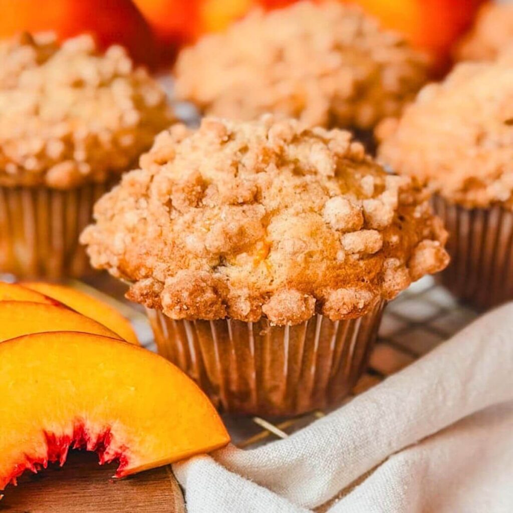 close up of a peach streusel muffin on a wire rack with sliced peaches next to the muffin.