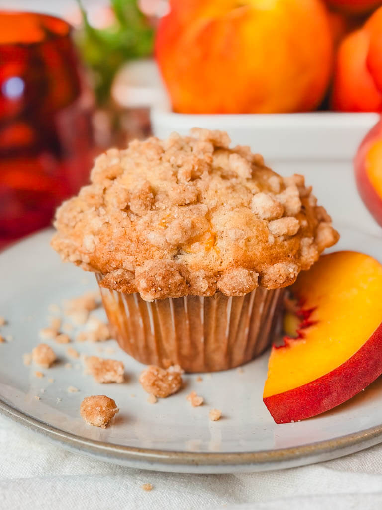 a peach muffin on a ceramic plate with streusel crumbs surrounding the muffin.
